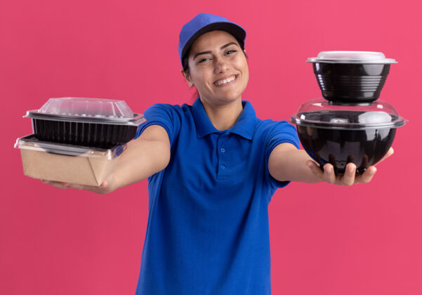 穿着面带微笑的年轻送货员身穿制服 头戴鸭舌帽 把食物容器放在隔离在粉红色墙上的前面制服抱着人