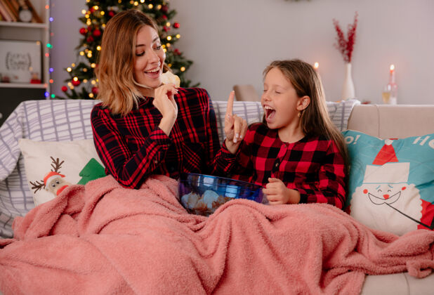 时间快乐的母亲拿着薯片和手势等着女儿闭上眼睛盖上毯子坐在沙发上 在家里享受圣诞节时光毯子圣诞节等待