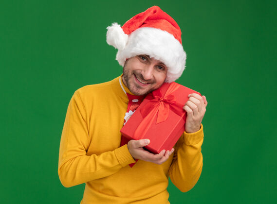 绿色快乐的年轻白种人戴着圣诞领带和帽子拿着礼包看着隔离在绿色背景下的相机年轻礼物男人