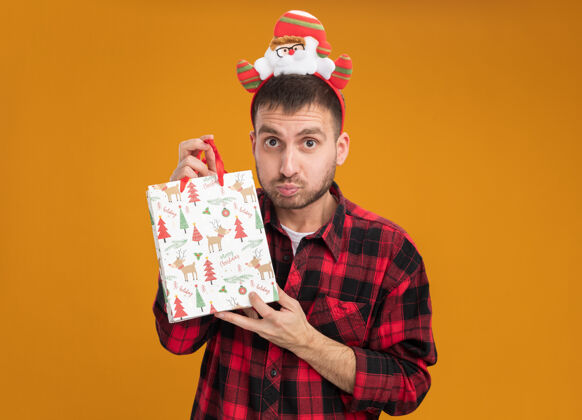 印象令人印象深刻的年轻白种人戴着圣诞老人的头带 拿着圣诞礼品袋 看着橙色背景上孤立的摄像机礼物圣诞老人复制