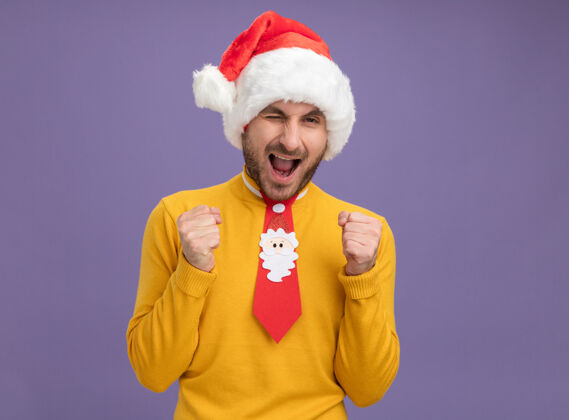 领带快乐的白人年轻人戴着圣诞帽打着领带看着摄像机眨眼做着“是”的手势孤立在紫色背景上圣诞快乐帽子男人
