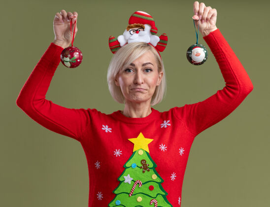 头带笨手笨脚的中年金发女人戴着圣诞老人的头带和圣诞毛衣举起圣诞饰品看着橄榄绿背景上孤立的相机无知加薪金发