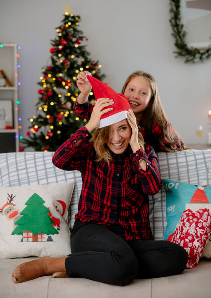 坐快乐的女儿把圣诞帽戴在妈妈头上 坐在沙发上 在家里享受圣诞时光快乐头圣诞老人