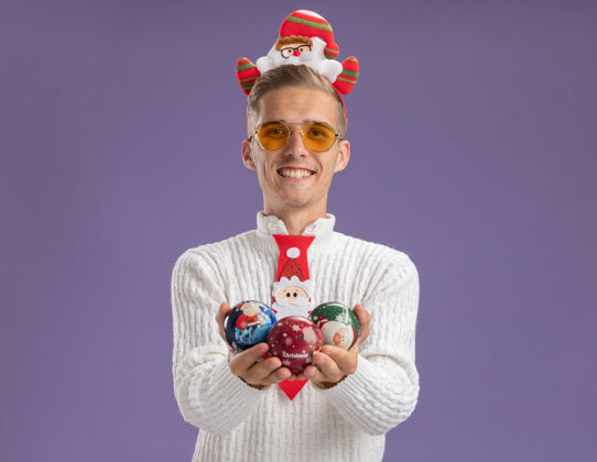 空间快乐的年轻帅哥戴着圣诞老人的头带和领带 戴着眼镜 拿着圣诞球饰品 看着隔离在紫色背景上的相机领带圣诞老人年轻人