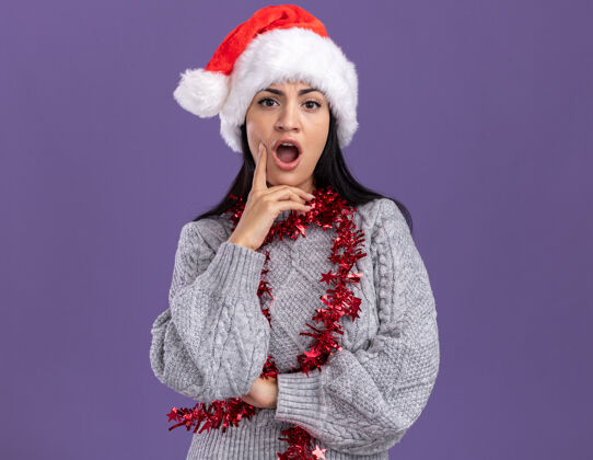 镀金令人印象深刻的年轻白人女孩戴着圣诞帽 脖子上戴着金箔花环 手放在下巴上 看着紫色背景上孤立的摄像机紫色女孩圣诞快乐