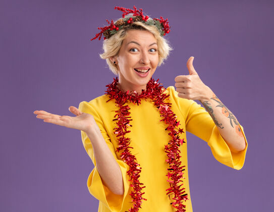空快乐的金发女郎戴着圣诞花环 脖子上戴着金属丝花环 看着镜头 在紫色背景下 她空手而起 拇指向上花环年轻周围