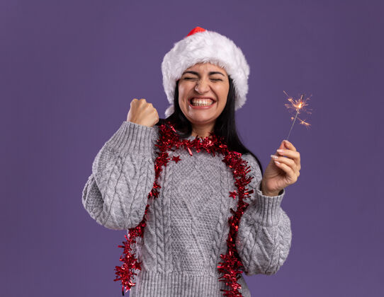 花环快乐的年轻白人女孩戴着圣诞帽 脖子上戴着金箔花环 手里拿着节日火花灯 做着“是”的手势 闭着眼睛 孤立地站在紫色的背景上年轻金属片圣诞快乐