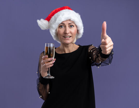 香槟快乐的中年金发女人戴着圣诞帽看着相机拿着一杯香槟在紫色背景上孤立地竖起大拇指复制举行中年
