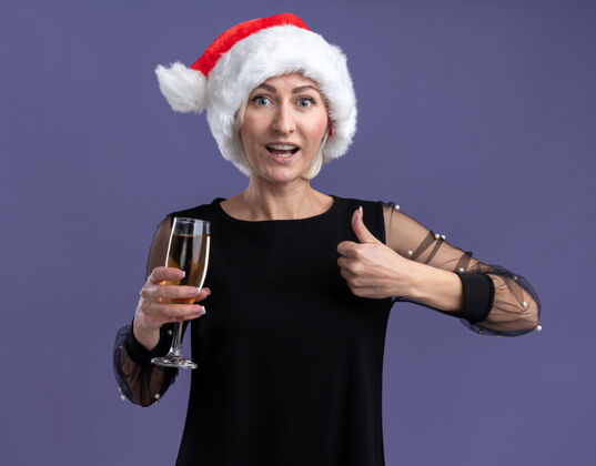 圣诞快乐令人印象深刻的中年金发女人戴着圣诞帽看着相机拿着一杯香槟在紫色背景上孤立地竖起大拇指帽子紫色中年