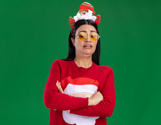 关闭恼怒的年轻白种女孩戴着圣诞老人的头巾和毛衣 戴着眼镜 站在绿色的背景下 以封闭的姿势俯视着与世隔绝头带圣诞老人复制