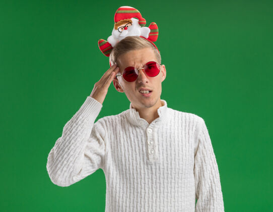 头带迷茫的年轻帅哥戴着圣诞老人的头带 戴着眼镜 一边看一边摸着绿色背景上孤立的头家伙年轻触摸