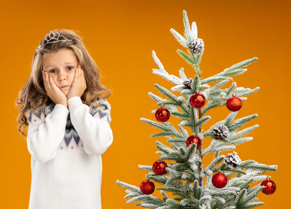 手关心的小女孩站在圣诞树旁 戴着头饰 脖子上戴着花环 手放在脸颊上 背景是橙色的头饰关心小