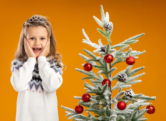 脸颊兴奋的小女孩站在圣诞树旁 戴着头饰 脖子上戴着花环 手放在脸颊上 背景是橙色的圣诞女孩站