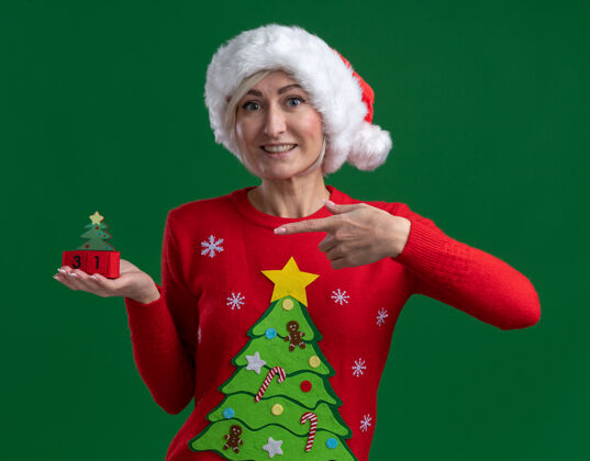 绿色快乐的中年金发女人戴着圣诞帽和毛衣拿着圣诞树玩具和日期看着相机隔离在绿色的背景玩具拿着中年