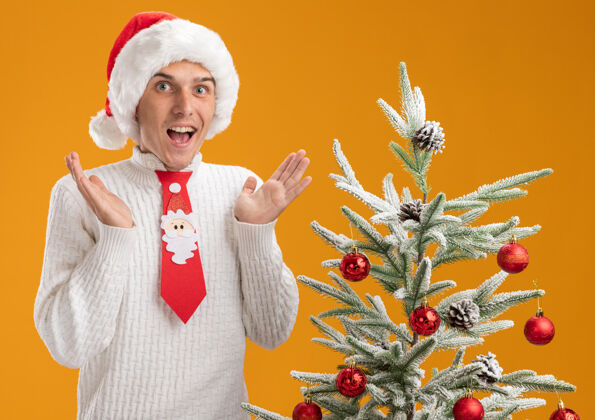 领带年轻帅哥戴着圣诞帽 打着圣诞老人的领带 站在装饰好的圣诞树旁 看着镜头 两手空空的 孤立在橙色的背景上手表演圣诞快乐