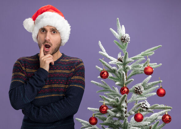 圣诞快乐令人印象深刻的年轻白种人戴着圣诞帽站在装饰过的圣诞树旁看着相机手放在下巴上孤立的紫色背景圣诞树印象手