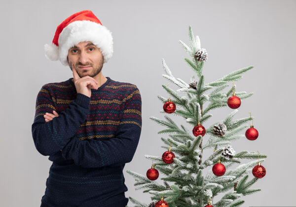圣诞戴着圣诞帽的可疑的白人年轻人站在圣诞树旁看着相机 手放在下巴上 在白色背景下保持隔离站白色手
