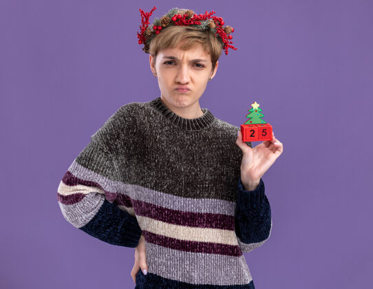 年轻皱眉的年轻漂亮女孩戴着圣诞花环手持圣诞树玩具和日期看着相机手放在腰上孤立在紫色背景上圣诞紫色手