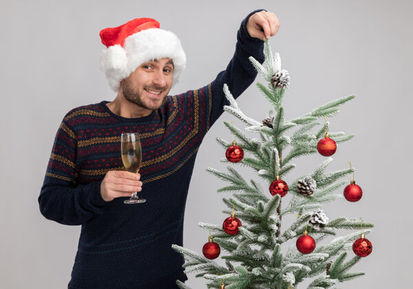 新年快乐的白人年轻人戴着圣诞帽 站在圣诞树旁 手里拿着一杯香槟 看着相机抓取隔离在白色背景上的树枝抓住快乐年轻