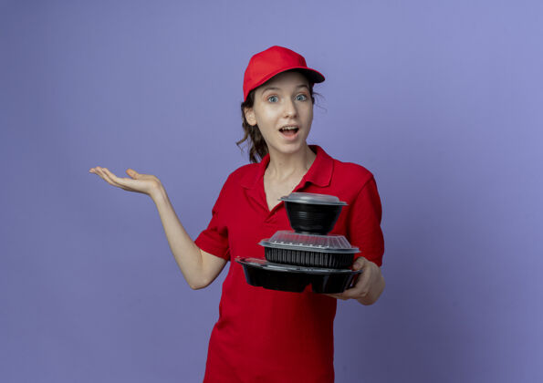 年轻令人印象深刻的年轻漂亮的送货女孩穿着红色制服和帽子举行食品容器显示空的手孤立在紫色背景与复制空间女孩漂亮持有