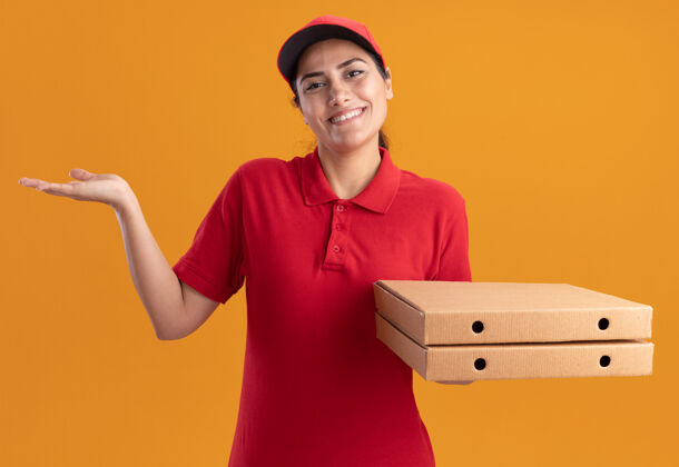 姿势微笑着的年轻送货女孩穿着制服 戴着帽子 手里拿着披萨盒 手被隔离在橙色的墙上盒子微笑女孩
