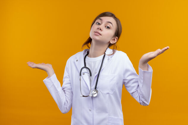 女性困惑的年轻女医生穿着医用长袍和听诊器 在孤立的橙色空间里空手而归穿医生手