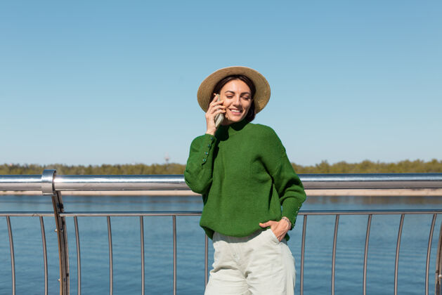 放松穿着绿色休闲毛衣 戴着帽子的时髦女人在户外的桥上 在温暖阳光下的夏日用手机聊天欢乐休闲人