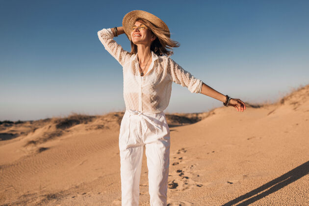 旅游时髦的微笑美丽的快乐女人穿着白色的衣服在沙漠中奔跑和跳跃在日落时戴着草帽微笑女人时尚