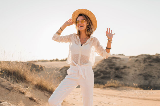 旅行时尚快乐的美丽微笑的女人在沙漠里摆姿势 穿着白色的衣服 戴着草帽和太阳镜在夕阳下非洲旅行者热