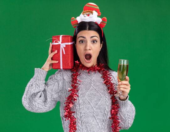 头带年轻的白人女孩戴着圣诞老人的头带 脖子上戴着金箔花环 肩上扛着礼包 喝着香槟 看着绿色背景上的相机年轻圣诞老人玻璃