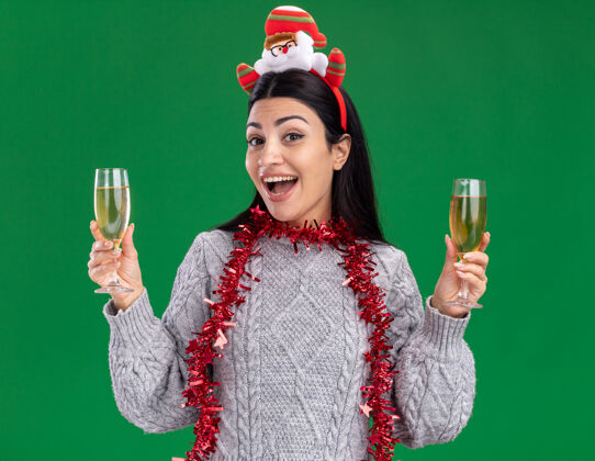 印象年轻的白人女孩戴着圣诞老人的头带 脖子上戴着金箔花环 手里拿着两杯香槟 看着隔离在绿色背景上的相机镀金两个香槟