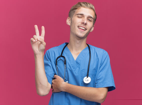 表情戴着医生制服的年轻男医生高兴地眨着眼睛 听诊器显示出和平的姿态 隔离在粉红色的墙上站立手势眨眼