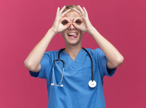 医生快乐的年轻男医生穿着医生制服 听诊器显示舌头和看手势隔离在粉红色的墙上年轻人快乐舌头