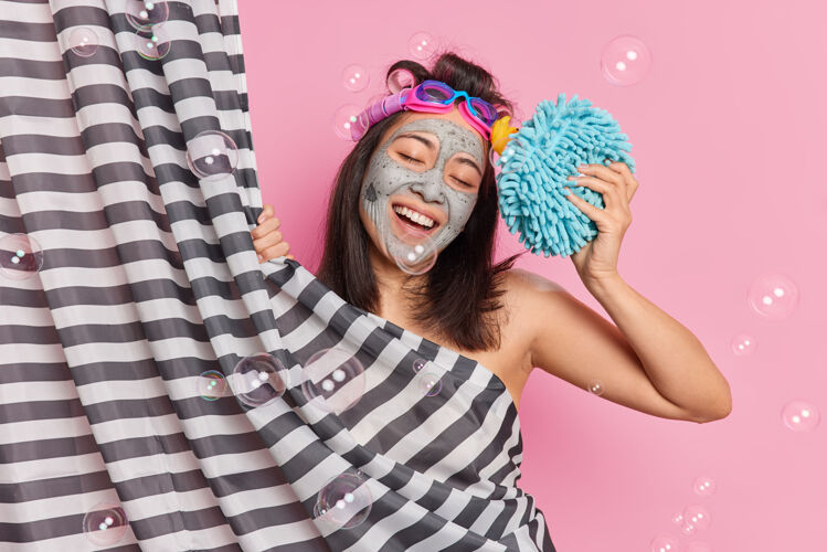 美女积极年轻的深色头发的亚洲女子微笑着快乐地歪着头在浴室洗澡享受卫生程序应用粘土面膜拿海绵洗身做发型满意肥皂放松