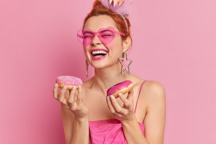 美味照片中快乐时尚的红发女人戴着时髦的太阳镜微笑着宽大地拿着美味的甜甜圈爱吃甜食年轻小吃牙齿