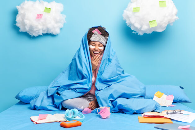 纸快乐的非洲裔美国女孩快乐地笑着有着乐观的心情裹着柔软的毯子在床上摆姿势准备考试垫休闲睡眠面罩