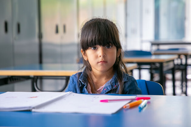 抄写本严肃的拉丁女孩坐在课桌旁 看着前面室内肖像小学生
