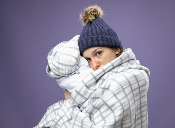 年轻向前看 年轻的生病的女孩穿着白色长袍 戴着冬天的帽子 围巾裹着格子呢 抱枕孤立地躺在紫色的床上疾病拥抱枕头