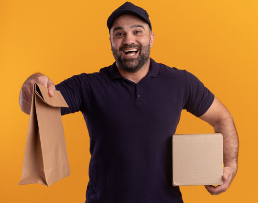 包装快乐的中年送货员穿着制服 戴着帽子 拿着盒子 拿着隔离在黄色墙上的纸食品包快乐拿着制服