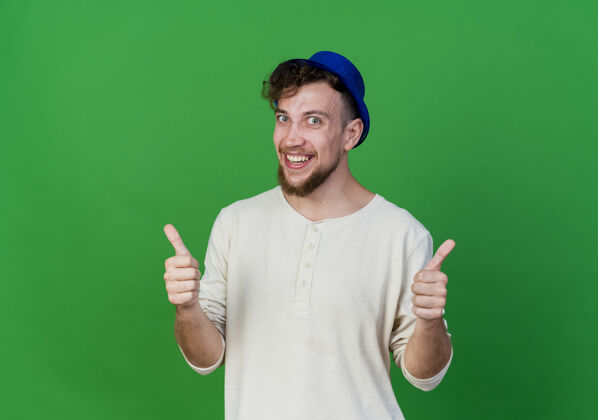 表演快乐的年轻英俊的斯拉夫党人戴着党的帽子 在前面显示拇指向上孤立的绿色墙壁与复制空间年轻人斯拉夫人站