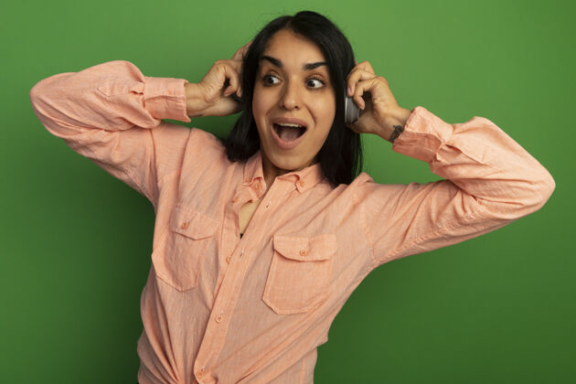 年轻惊讶地看着身边年轻漂亮的女孩穿着粉色t恤 把耳机隔离在绿色的墙上女孩耳机壁板