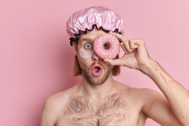 补丁带着鲍勃发型的震惊的成年男子用甜甜圈遮住眼睛盯着镜头 在粉色背景下赤膊上阵身体奇迹甜甜圈