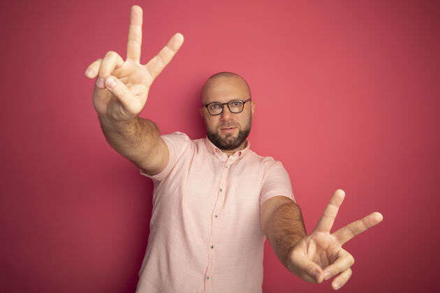 中年高兴的中年秃头男子穿着粉色t恤 戴着眼镜 显示出和平的姿态 孤立在粉色上男人眼镜姿态