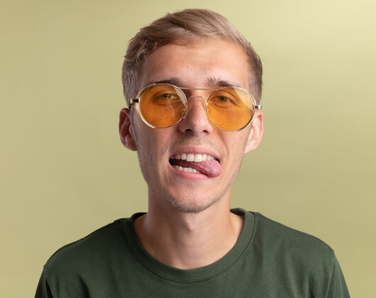 橄榄快乐的年轻帅哥穿着绿色衬衫 戴着眼镜 在橄榄绿的墙上露出孤立的舌头小伙子秀帅哥