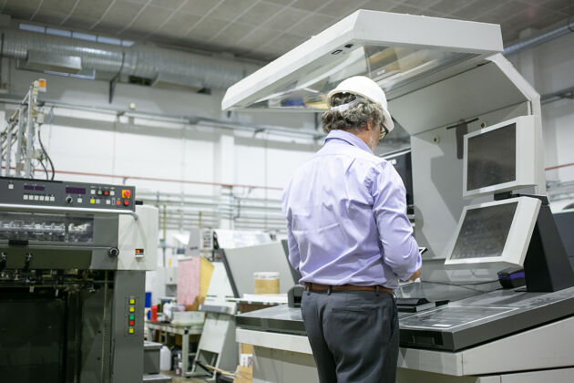 生产白发苍苍的男性工厂工程师戴着安全帽 戴着眼镜站在工业机器前 用的是数码设备安全帽平板电脑眼镜