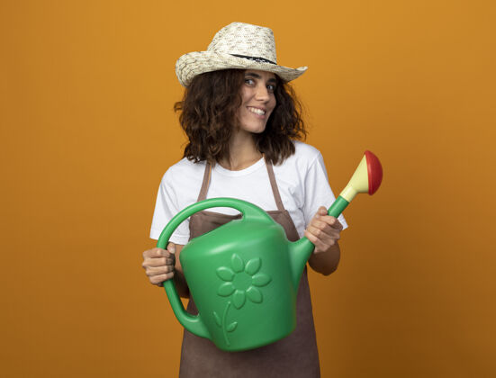 女人微笑着的年轻女园丁 穿着制服 戴着园艺帽 手里拿着浇水罐穿浇水举行