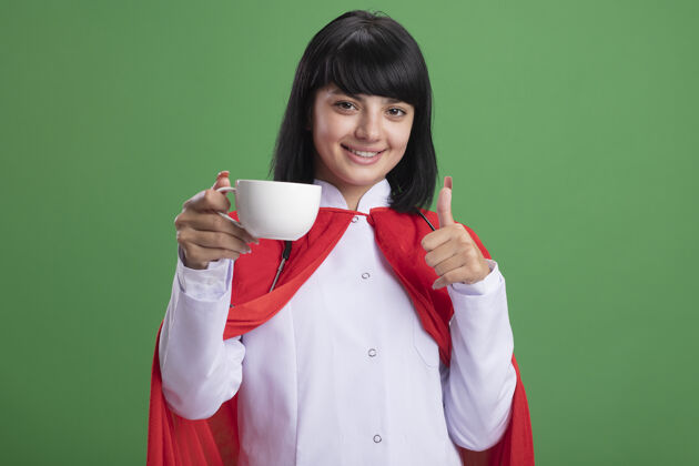 超级英雄微笑着的年轻超级英雄女孩戴着听诊器 穿着医用长袍 披着斗篷 手里拿着一杯茶 在绿色的草地上孤立地竖起大拇指显示举行杯子