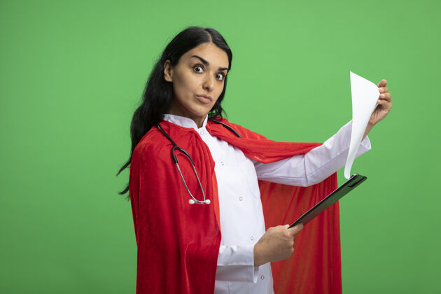 绿色惊讶的年轻超级英雄女孩穿着医用长袍 手持听诊器 笔直地看着前方 在绿色的剪贴板上翻阅穿医疗举行