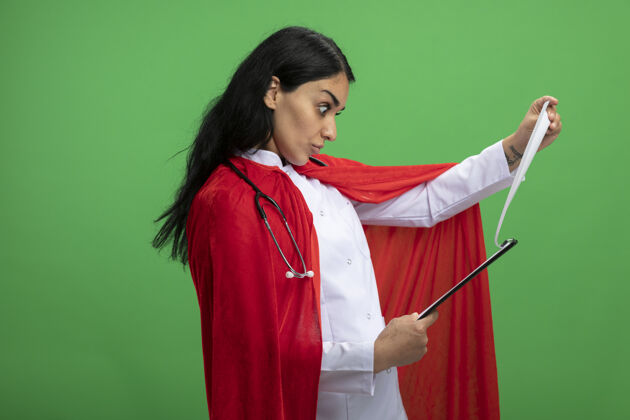 穿惊讶的年轻超级英雄女孩穿着医用长袍 手持听诊器 在绿色的剪贴板上翻来翻去长袍惊喜超级英雄