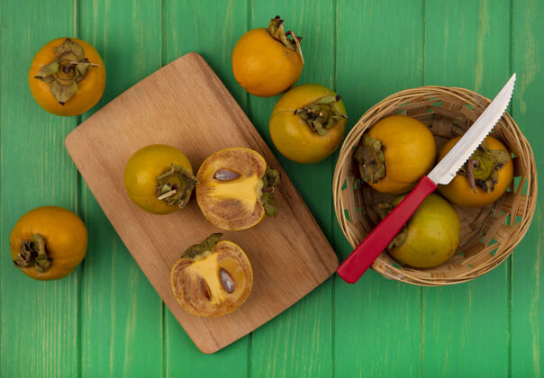 食物新鲜柿子水果的顶视图在一个桶上 在一张绿色的木桌上 用刀把柿子水果切成两半放在一个木制的厨房板上一半木头绿色